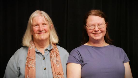 Barbara Sjoholm and Amy Swanson King pose in front of a black curtain in a studio.