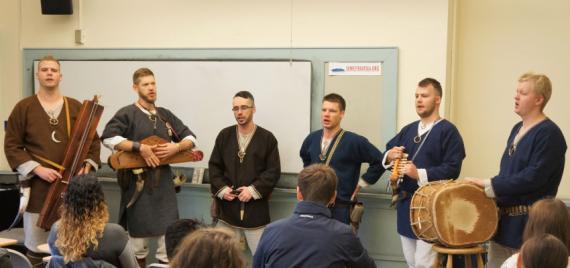 Vilkači performs in a UW classroom. Six band members wear medieval clothing and play traditional instruments while they sing.