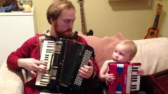 Linus plays accordion on a couch with his child who plays a child's accordion.