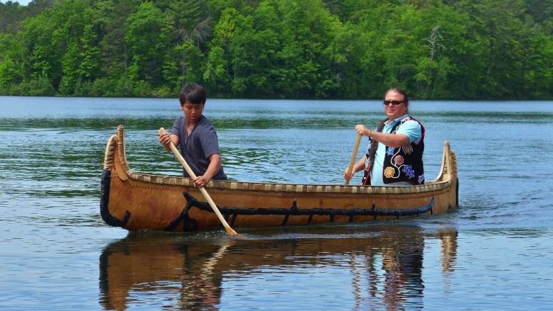 An Ojibwe elder and youth paddle a birchbark canoe together on a lake.
