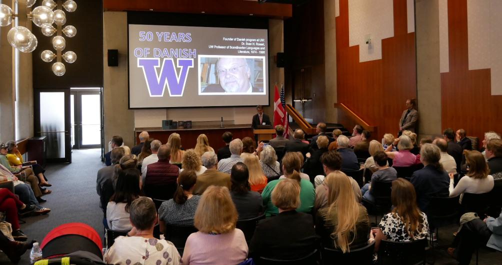 Faculty, staff, and alumni sit attentively to listen to opening remarks in a room at the university. A slide projector shows an image of Sven Rossel's face and reads, "50 years of Danish at UW."
