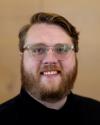 Ethan Bjelland Hagberg has clear-framed glasses, a black shirt, dark blonde hair and a beard, and they are smiling at the camera in front of a light wooden background.