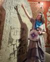 Lauren stands in front of a rune stone and point to the top. They are wearing a grey dress with a blue bandana. 