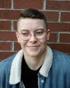 Subject has short brown hair, round glasses, a jean jacket with a fur lining and stands smiling in front of a brick wall.