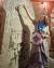 Lauren stands in front of a rune stone and point to the top. They are wearing a grey dress with a blue bandana. 