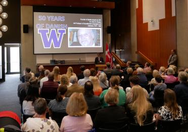 Faculty, staff, and alumni sit attentively to listen to opening remarks in a room at the university. A slide projector shows an image of Sven Rossel's face and reads, "50 years of Danish at UW."