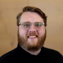 Ethan Bjelland Hagberg has clear-framed glasses, a black shirt, dark blonde hair and a beard, and they are smiling at the camera in front of a light wooden background.
