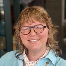 Headshot of Olivia Noble Gunn. Blue dress, green glasses, long gold chain around her neck and a small scarf in cream with red squiggles