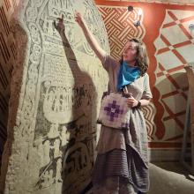 Lauren stands in front of a rune stone and point to the top. They are wearing a grey dress with a blue bandana. 